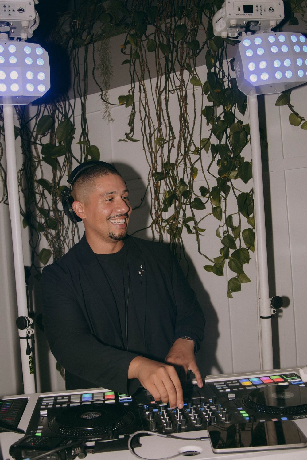 Andrew smiling behind the DJ booth with headphones on, white moving-head lights and hanging greenery behind him