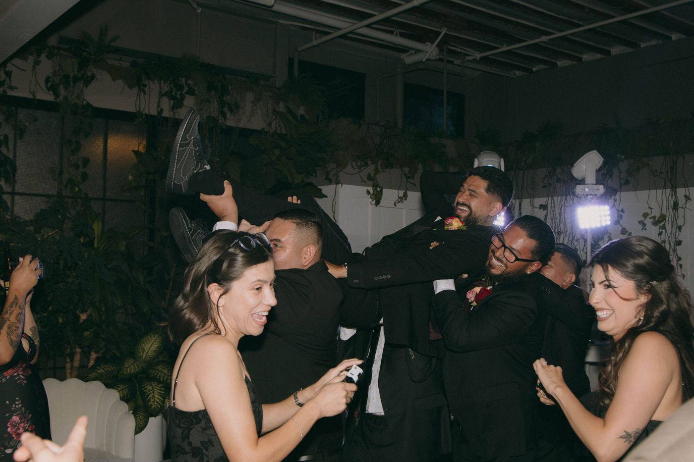 A man in a black suit is lifted into the air by friends on the dance floor, everyone laughing